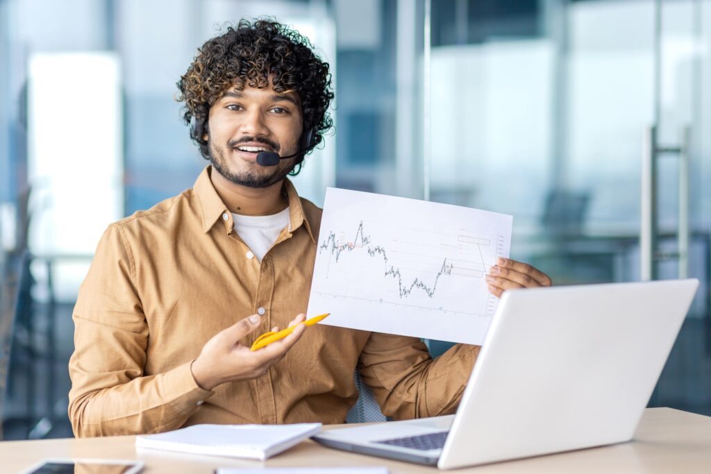 Man in headset learning forex trading, pointing pen at paper with charts in front of laptop.