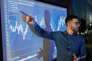 Business coach using a pen to highlight trading tools for beginners on a projector displaying market data.