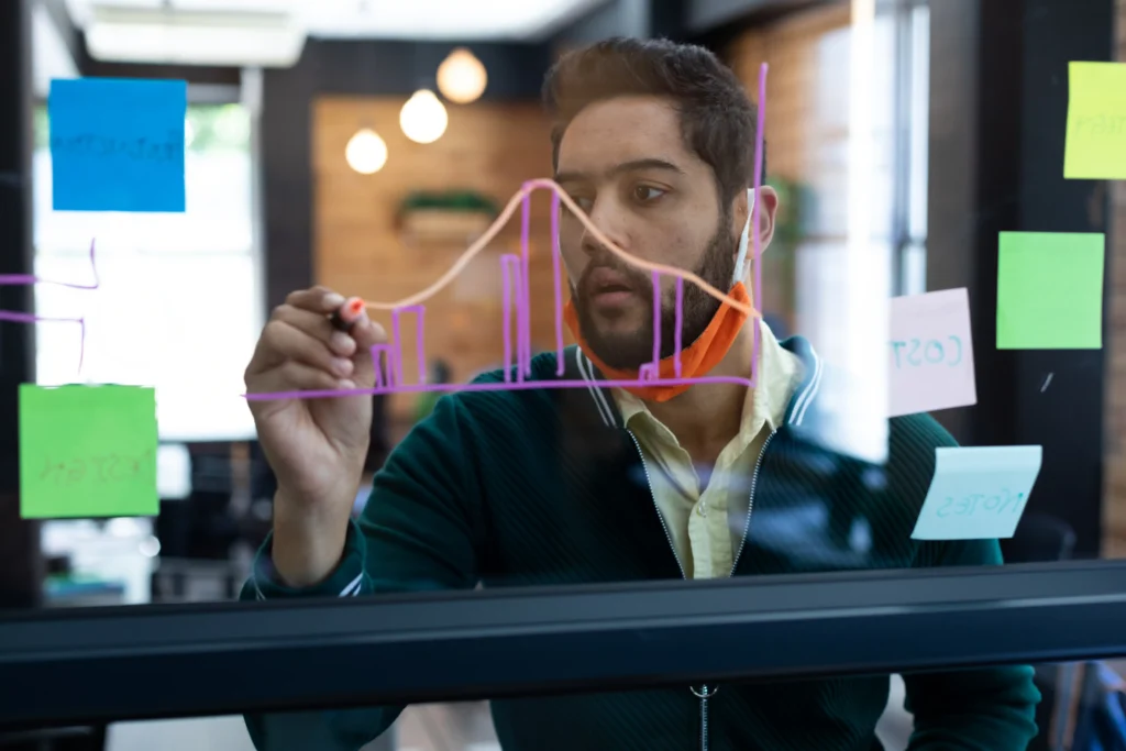 Man drawing financial chart with moving averages on transparent board in a modern office setting.