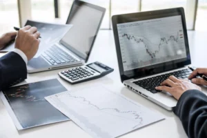 Traders reviewing forex charts on laptops with financial documents comparing prop firm accounts vs personal trading accounts.