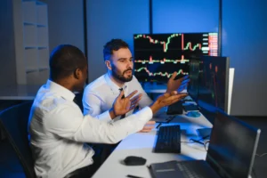 Two traders in front of multiple screens showing candlestick charts, engaged in market strategy discussion.