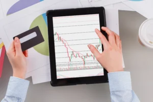 Person holding a tablet showing candlestick trading chart with a card in hand, representing transaction cost in trading.