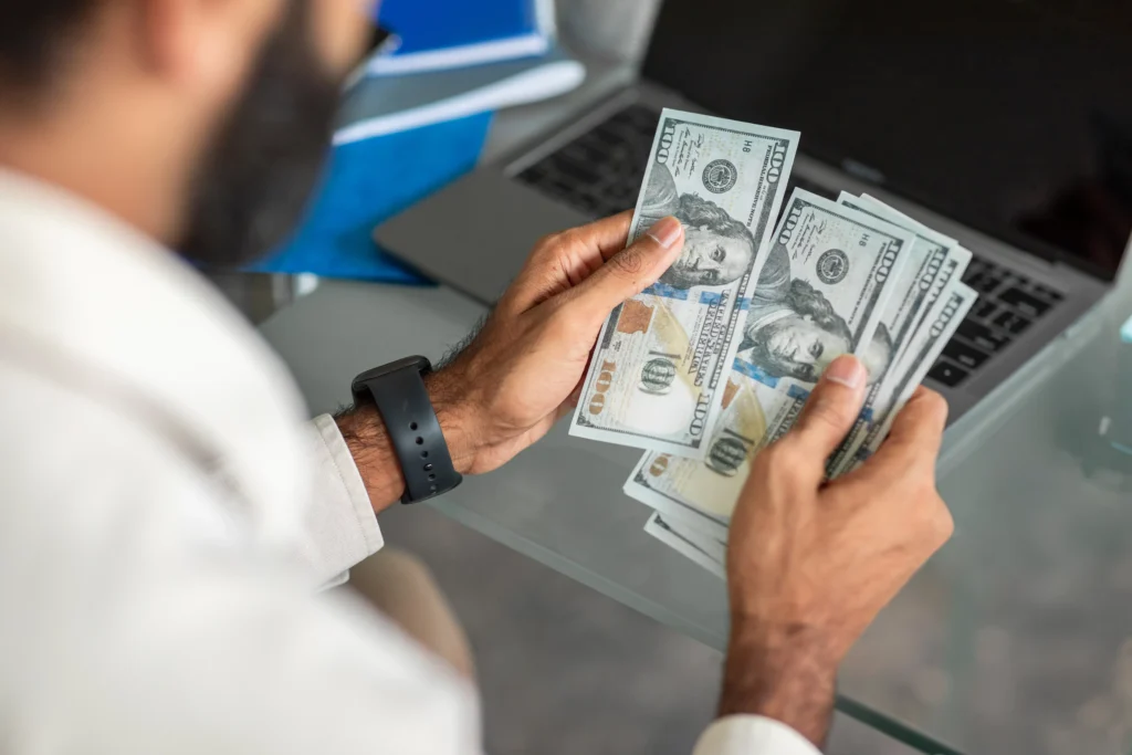 Person holding US dollars in hand near a laptop, representing trading capital and risk management for beginner traders.
