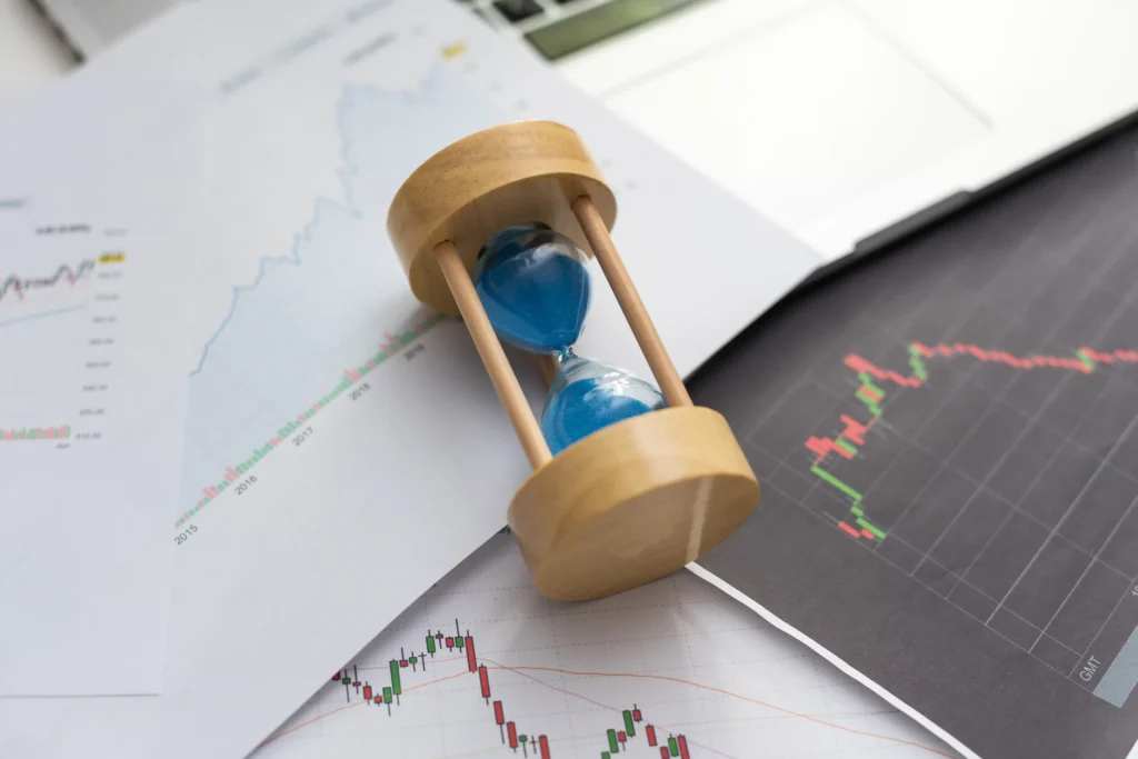 Wooden hourglass with blue sand on trading charts showing market predictions and price trends.