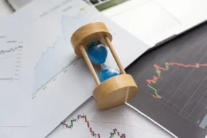 Wooden hourglass with blue sand on trading charts showing market predictions and price trends.