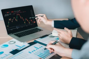 Two traders analyzing charts and data on a laptop, discussing Trading Terminology 2025 with forex and stock market trends.