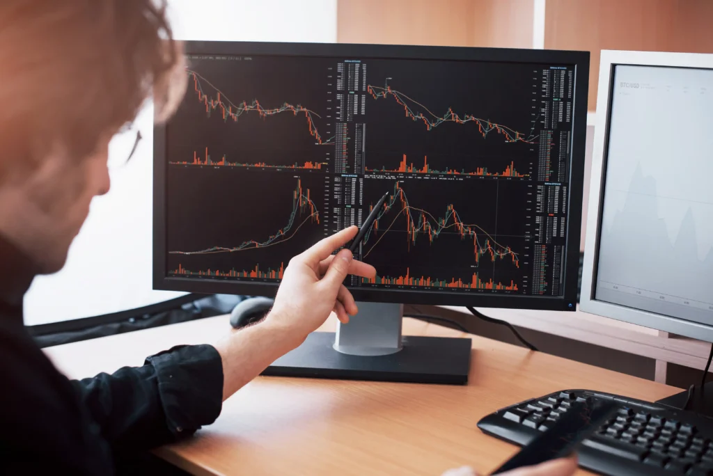 Trader pointing at multiple trading charts on a monitor.