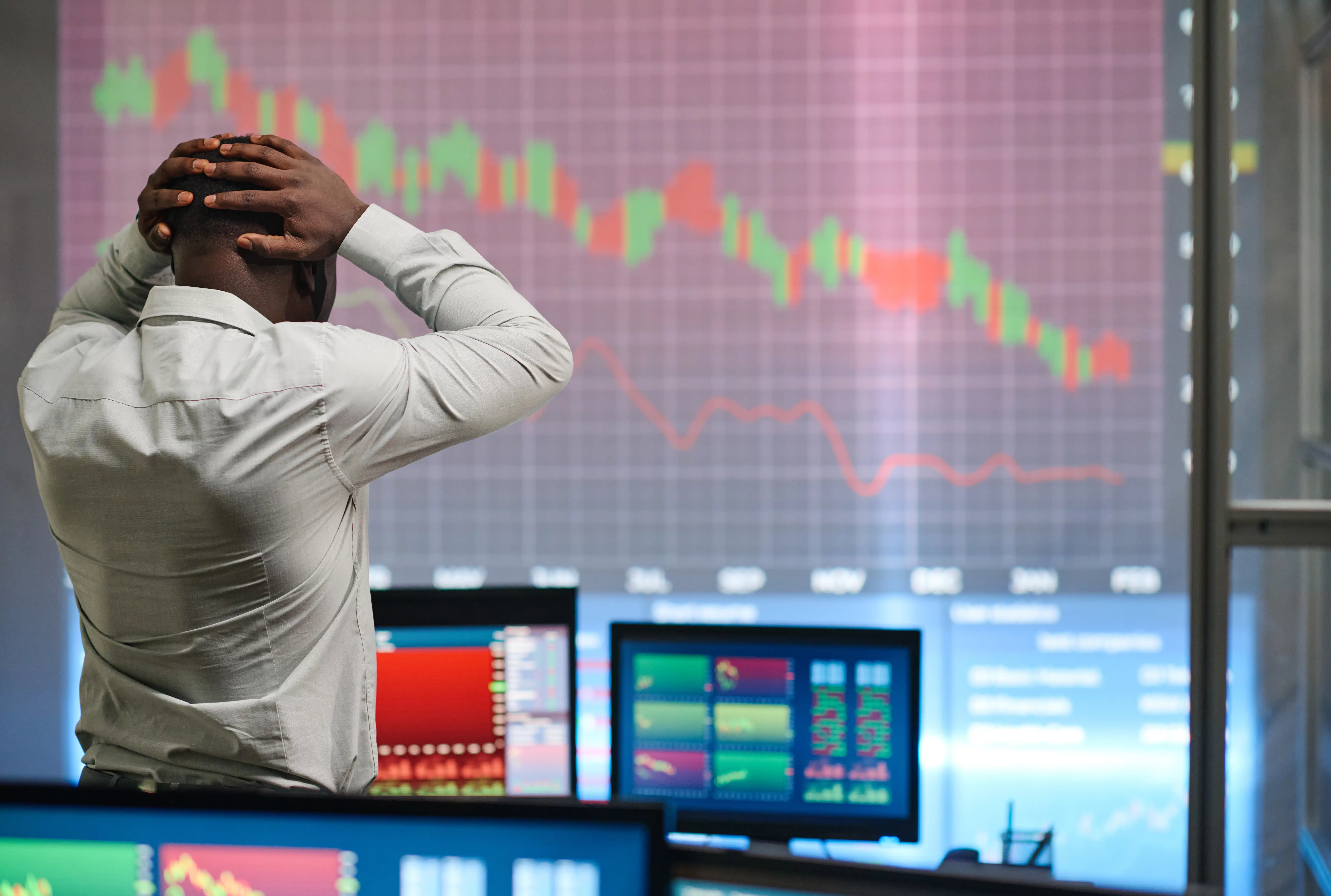 Trader holding head in frustration while watching falling stock and forex charts on multiple monitors.
