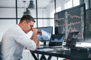 Stressed stock trader sitting at desk with multiple monitors showing volatile market charts.