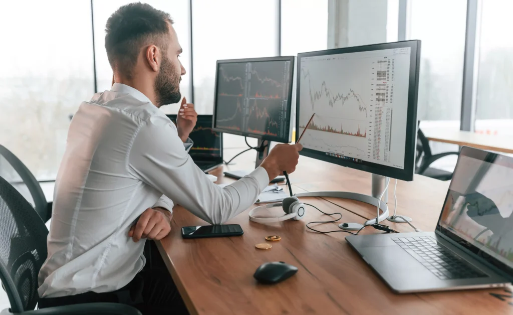 Man analyzing stock charts on dual monitors while discussing day trading tips for beginners in a modern office setting.