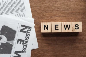 7 Smart Ways to Take Advantage of Forex News shown with wooden cubes spelling NEWS beside business newspapers on a wooden desk.