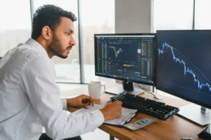 Focused trader analyzing live market charts on dual monitors while taking notes in a journal.