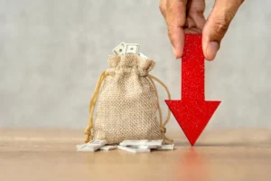 A burlap money bag with dollar bills spilling out beside a red downward arrow symbolizing currency devaluation.