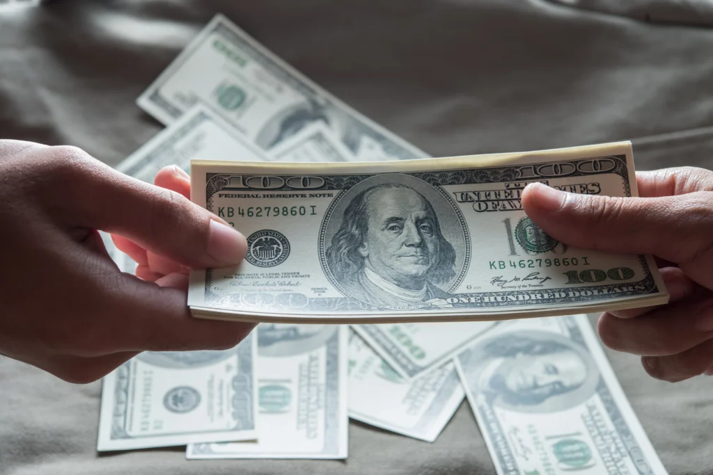 Two people exchange U.S. hundred-dollar bills, symbolizing global trade and the dominance of the U.S. dollar in international markets.