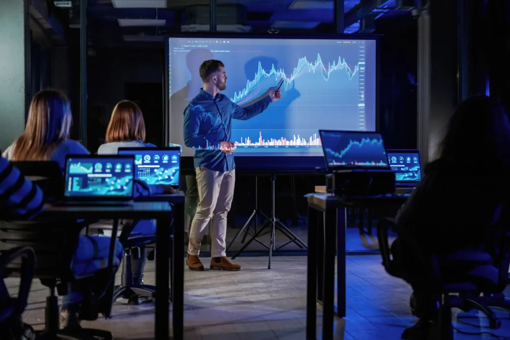A financial analyst presenting cross-asset forecasting data on a large screen to traders during a market analysis session.