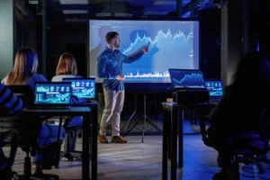 A financial analyst presenting cross-asset forecasting data on a large screen to traders during a market analysis session.