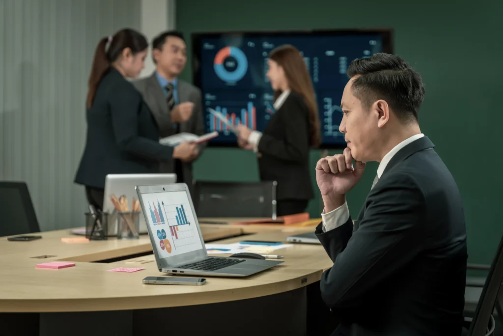 Asian businessman analyzing market charts on his laptop while colleagues discuss trading performance in the background, representing Telegram and Discord trading strategy planning.