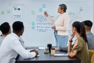 A professional team in a meeting room listens to a woman explaining strategy points on a whiteboard filled with business notes and planning diagrams.