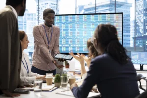 A group of people in a modern office discussing financial charts displayed on a large screen, while one person presents data using a tablet.