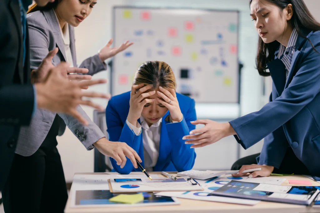Stressed businesswoman surrounded by colleagues during a tense discussion, representing holiday trading mistakes and emotional trading pressure in forex markets.