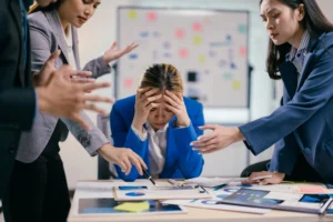 Stressed businesswoman surrounded by colleagues during a tense discussion, representing holiday trading mistakes and emotional trading pressure in forex markets.