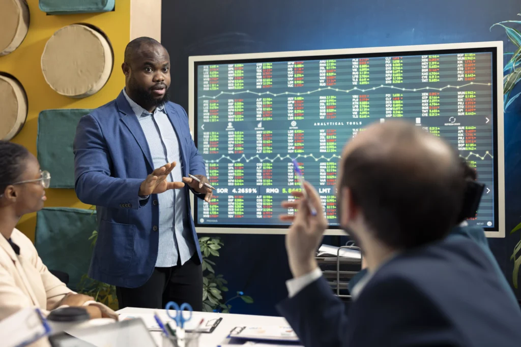 Financial expert explaining stock trends on screen to colleagues while discussing Trading Consistency.