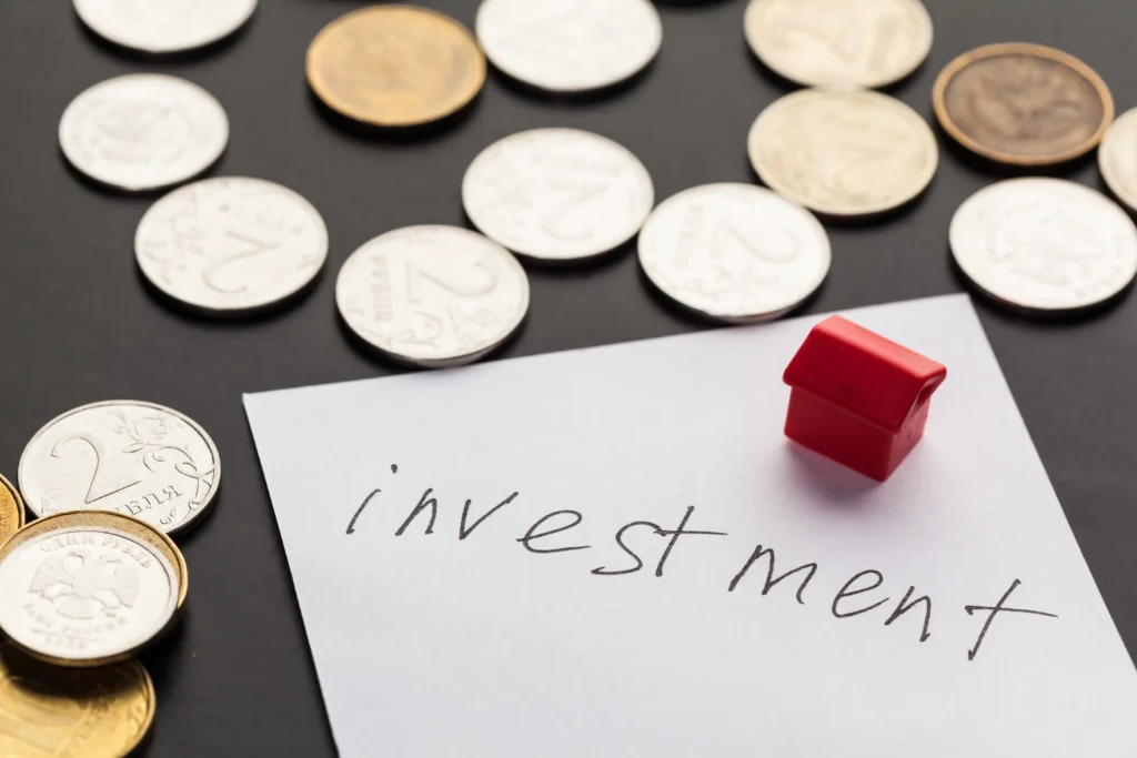 Coins scattered on a dark surface with a red miniature house and a paper labeled “investment,” symbolizing the concept of choosing the Right Investment Product 2026.